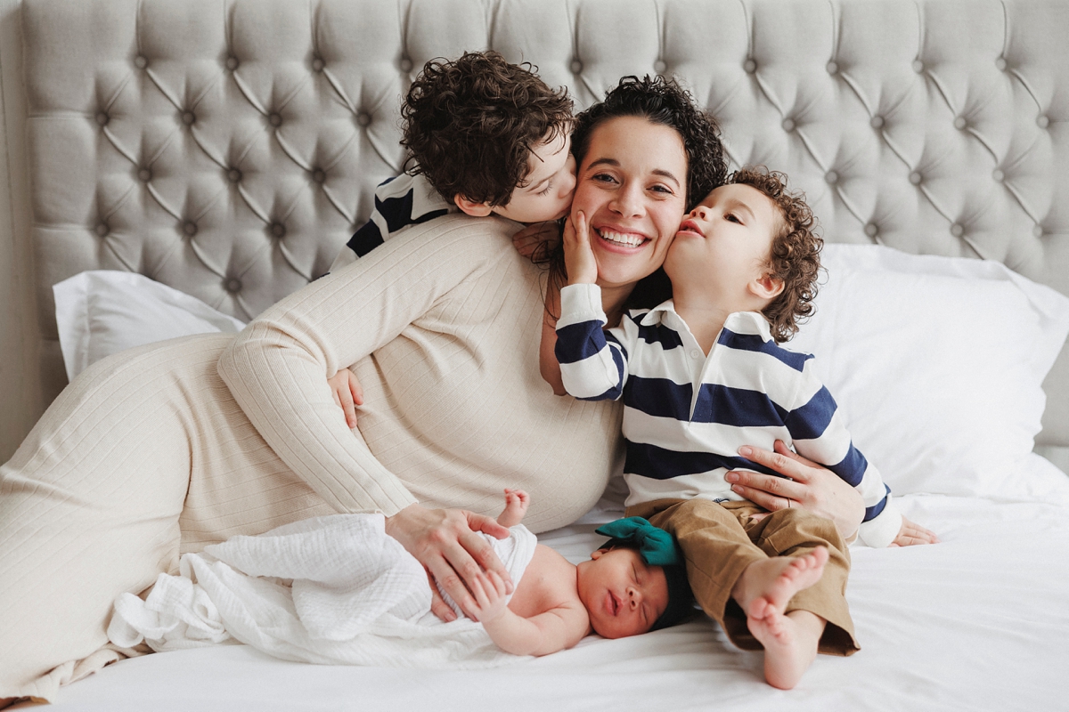 family photo in a chicago photo studio of two boys kissing mom and newborn on a white bed