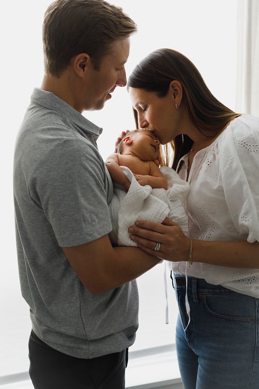 mom kisses babies forehead near a window during a newborn photoshoot in her home in chicago, IL