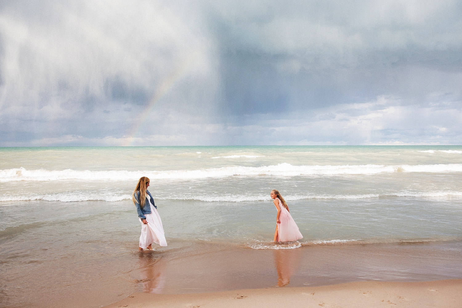 a mother and daughter stand on the beach under a rainbow togetehr