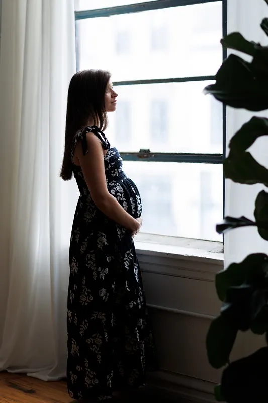 a pregnant woman holds her belly in a floral maxi dress against a window
