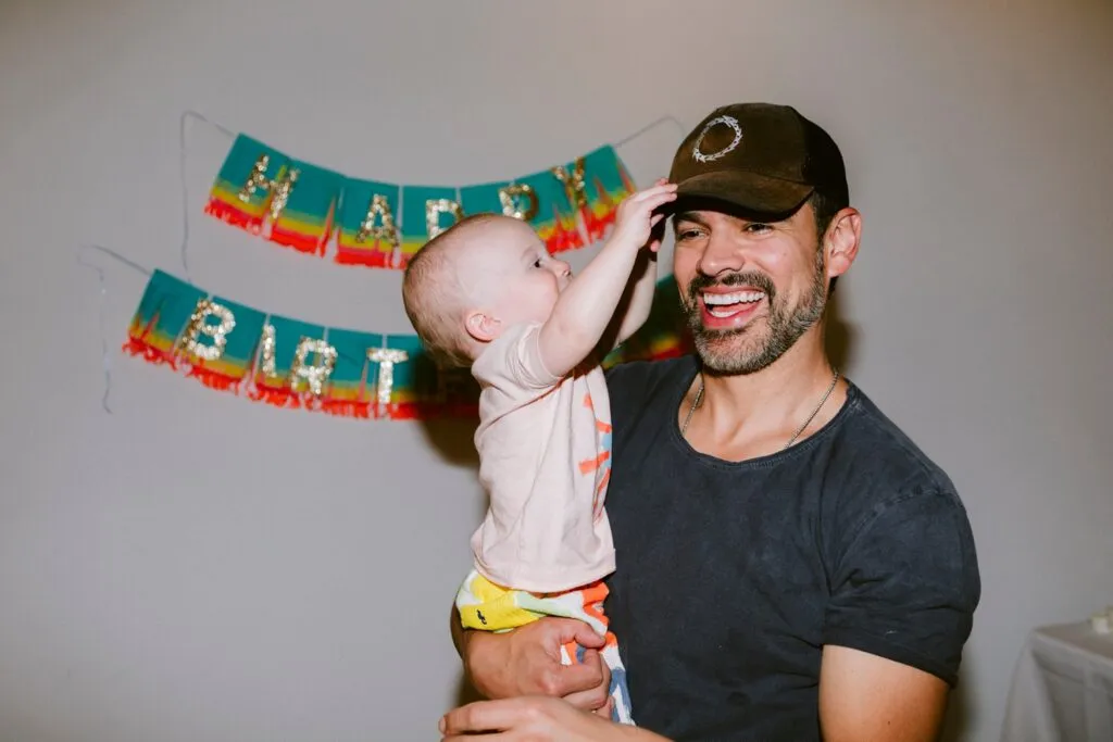 a man holds a baby at a first birthday party as he plays with his hat